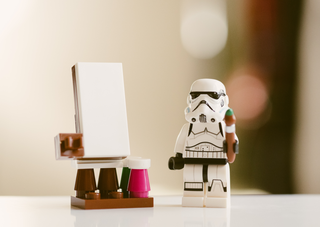 A Stormtrooper Lego figure holding a brush. The figure is standing in front of an easel and a blank canvas surrounded by paint.