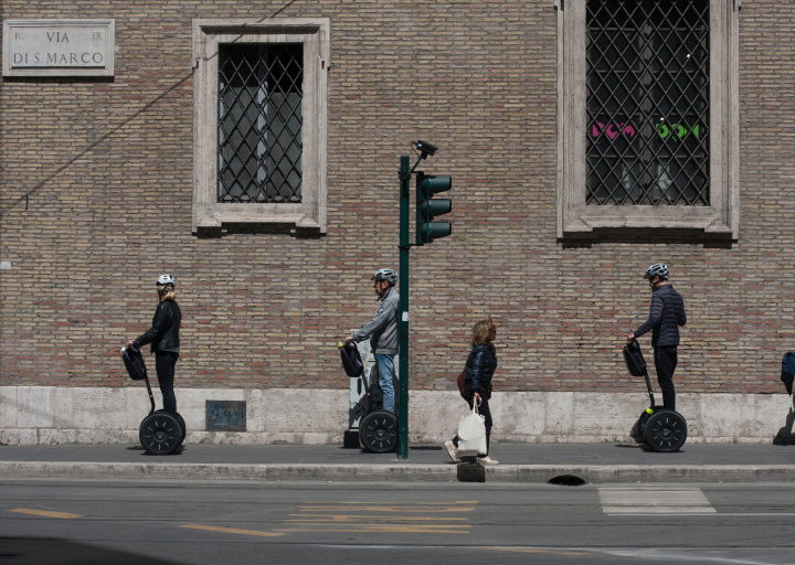 Three peaople riding Segway in a city. There is also a pedestrian walking in the opposite direction and making space for the Segways as there isn't much space on the pavement.