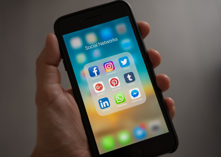 A hand holiding a phone with a "Social Networks" folder open. There are a Facebook icon, Instagram icon, Twitter, Google+, Pinterest, Twitch, LinkedIn, WhatsApp, and Messenger.