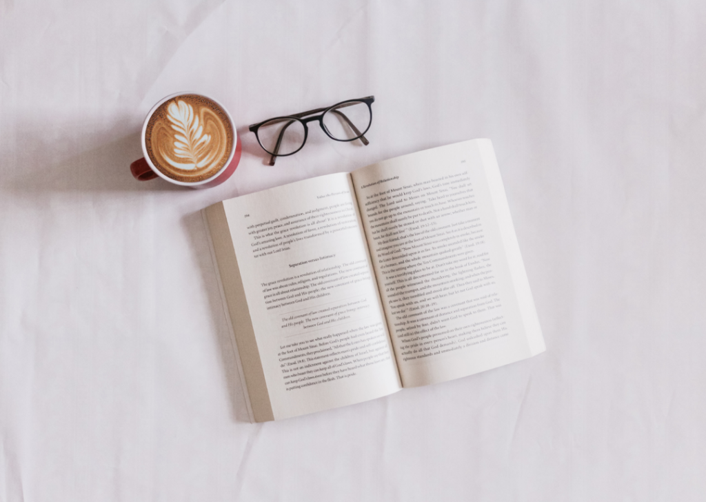 An open book, a cup of creamy cappuccino and a pair of reading glasses on a bed sheet.